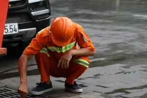 A street worker cleans a drainage system on a wet road after rainfall using a Canon EOS camera.